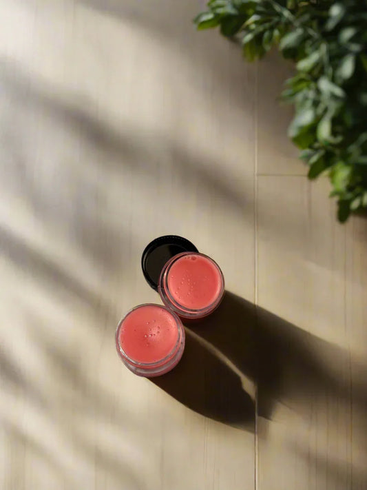 Two glass jars of pink henna balm on a sunlit wooden surface with green leaves nearby