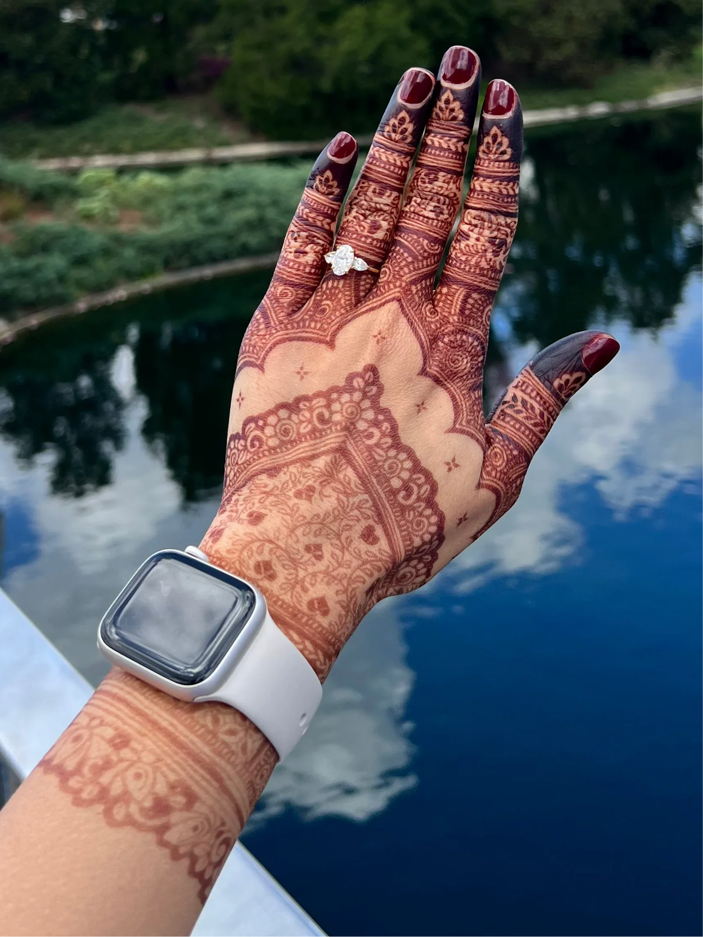 Hand with intricate henna artwork, diamond ring, smart watch, and outdoor lake background