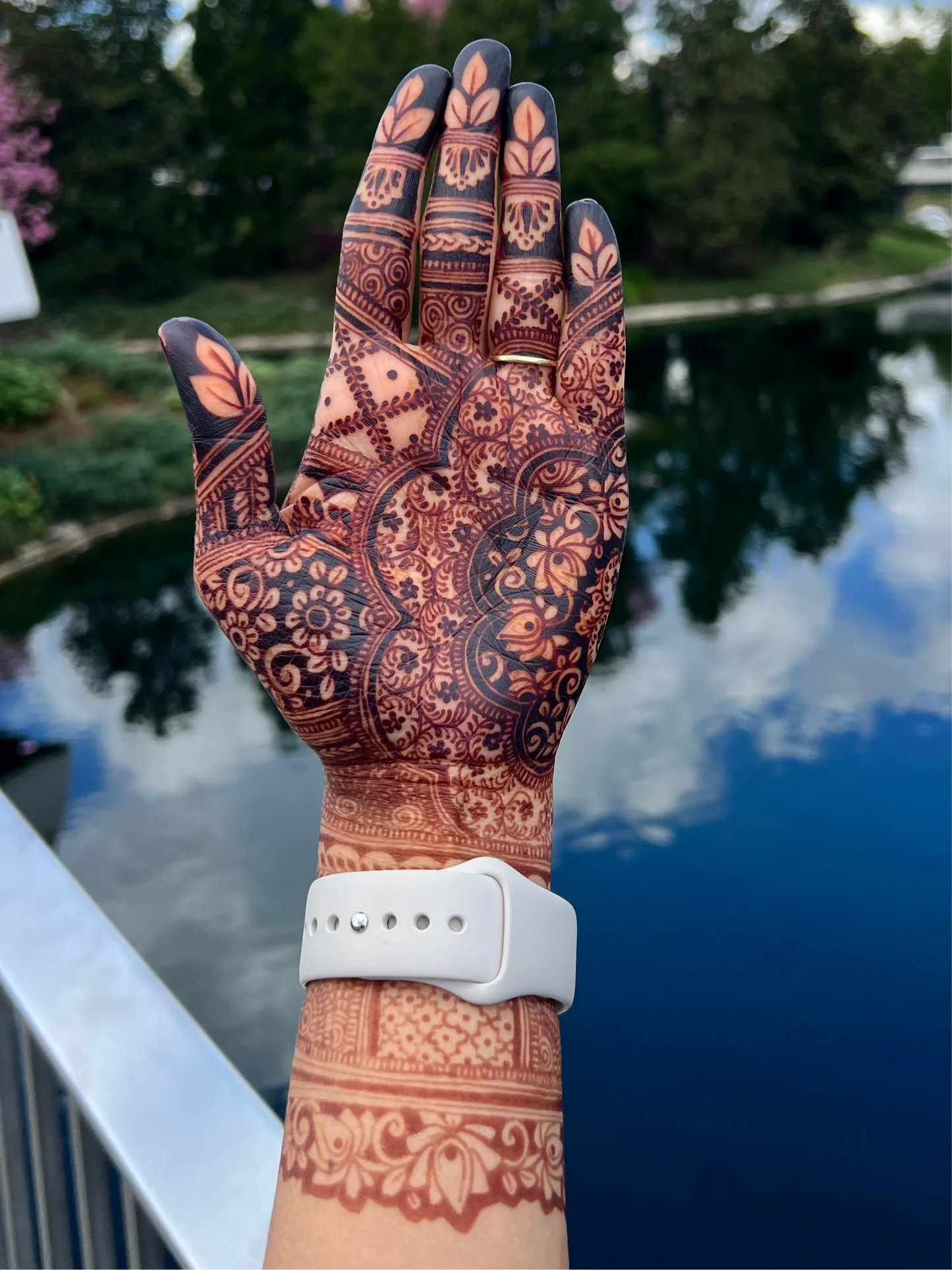 Hand with intricate floral henna artwork, white watch, near water and greenery outdoors