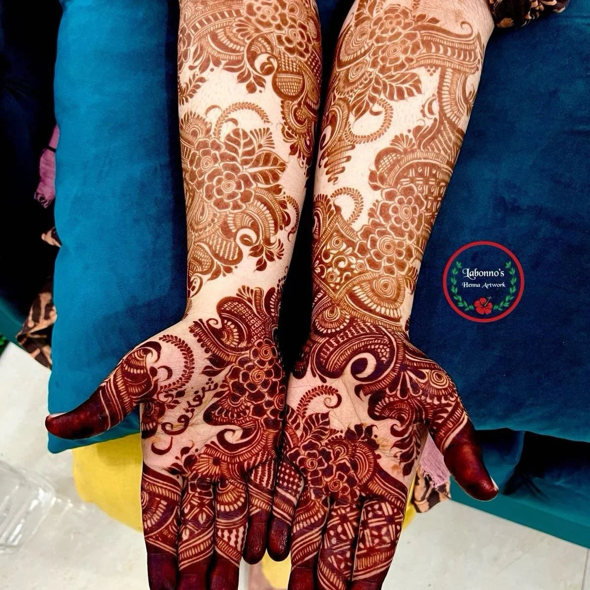 Intricate floral and paisley henna designs on both hands against a blue cushion background
