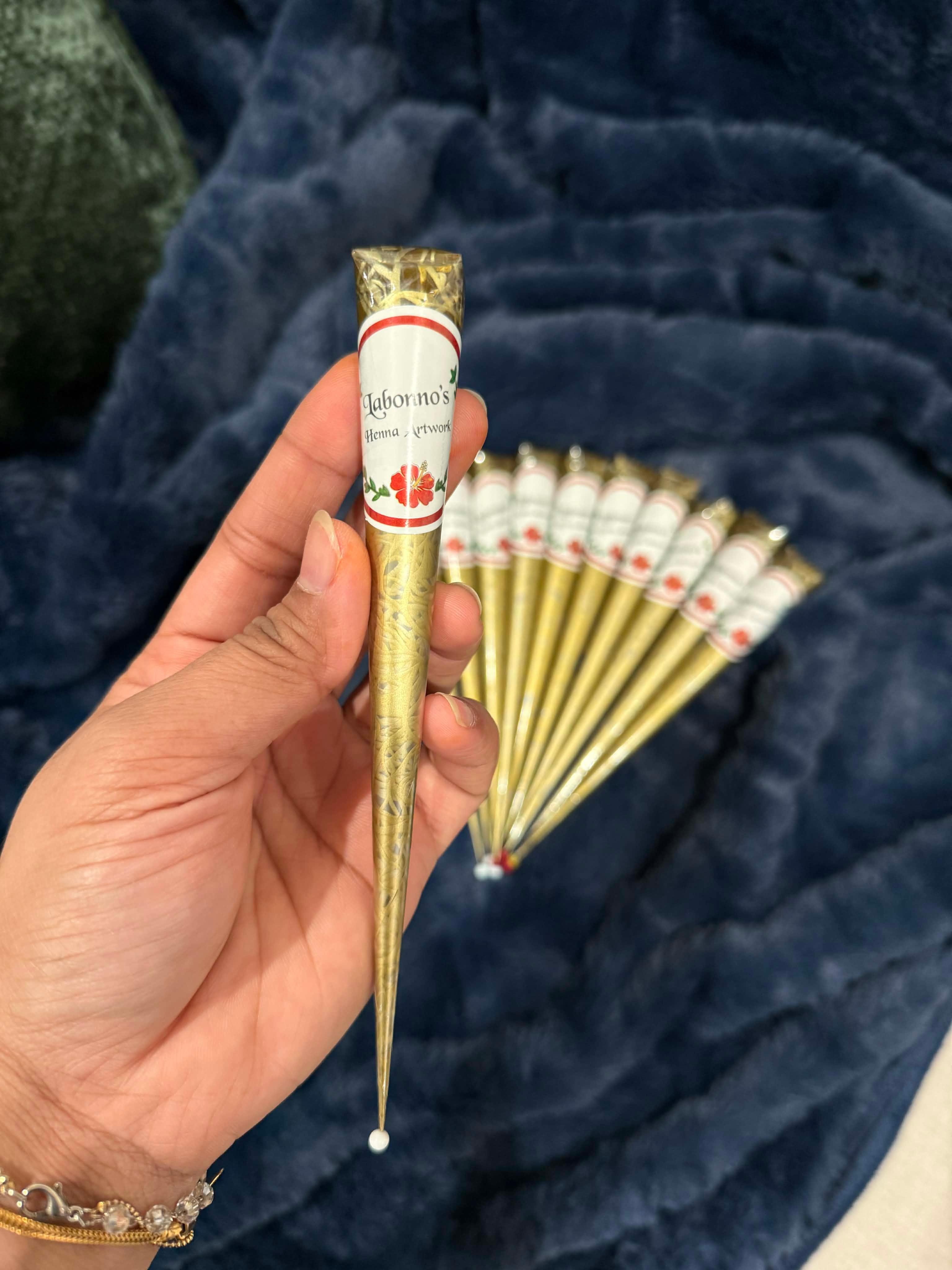 Hand holding gold henna cone with label, several henna cones on blue fabric background